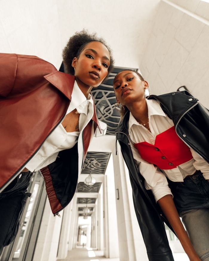 Two stylish women in modern attire pose confidently in an architectural corridor, showcasing bold fashion choices.